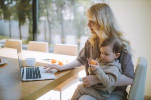 Busy mom holding baby while working on laptop at large table in the sunlight. Therapy in St. Petersburg, FL can help you cope with anxiety, depression, stress and overwhelm. Get started with online therapy in Florida with a St. Pete therapist here!